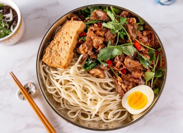 Bowl of noodles with spicy stir-fried beef, cilantro, tofu and a soft egg