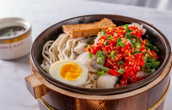 Spicy fish fillet mixed noodles with chopped chili, soft egg and tofu in a wooden bowl